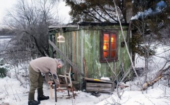 A lone man restoring an old wooden cabin in a snowy forest — day 5 of his wilderness retreat, carrying a broken chair back into the cabin