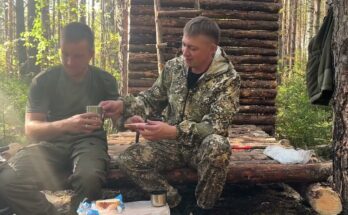 Two friends building a wooden house by hand — framing, saws, timber, and teamwork under a clear sky.